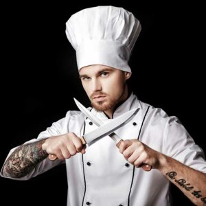 Young bearded man chef In white uniform holds Two knives on black background